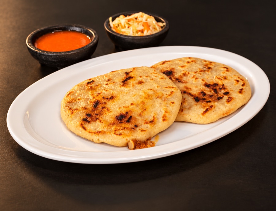 Two griddled pupusas on an oval plate with salsa and curtido in small black bowls.
