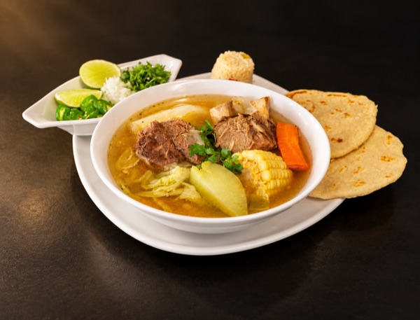 A bowl of clear soup with meat, corn, carrot, chayote and cabbage, with rice, lime, onion, cilantro and tortillas.