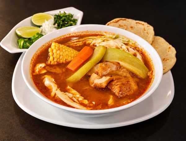A bowl of red broth soup with chicken, corn, carrot, chayote and cabbage, with lime, onion, cilantro and tortillas.
