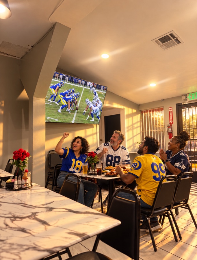 Fans in Rams and Cowboys jerseys cheering at the screen over plates and drinks; a staged promotional scene