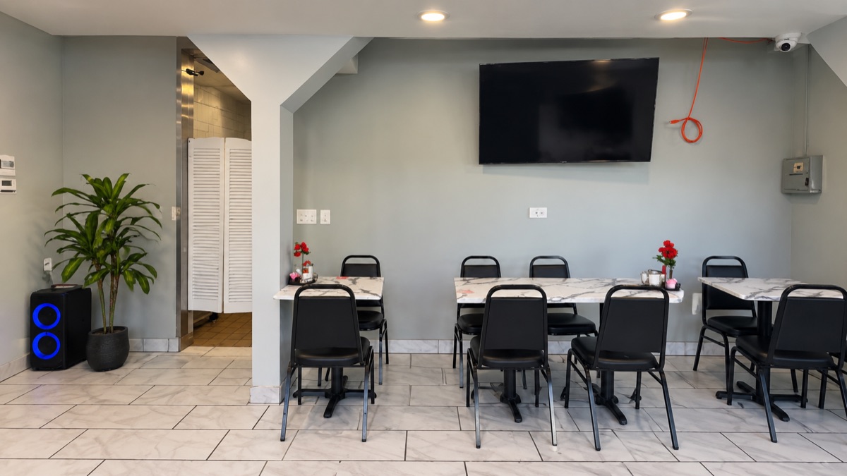 The dining room from the centre: grey walls, white marble tables with black chairs, a wall-mounted television and a tall plant beside the restroom doorway.